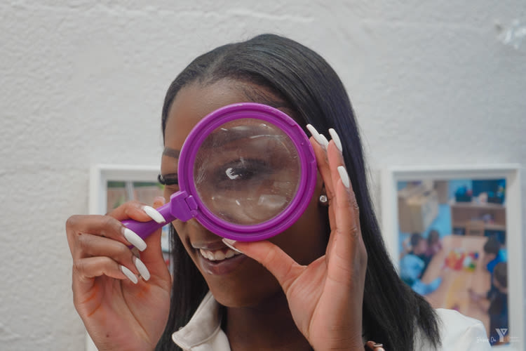 A close-up of an educator in a white blouse with dark hair looking through a purple magnifying glass.