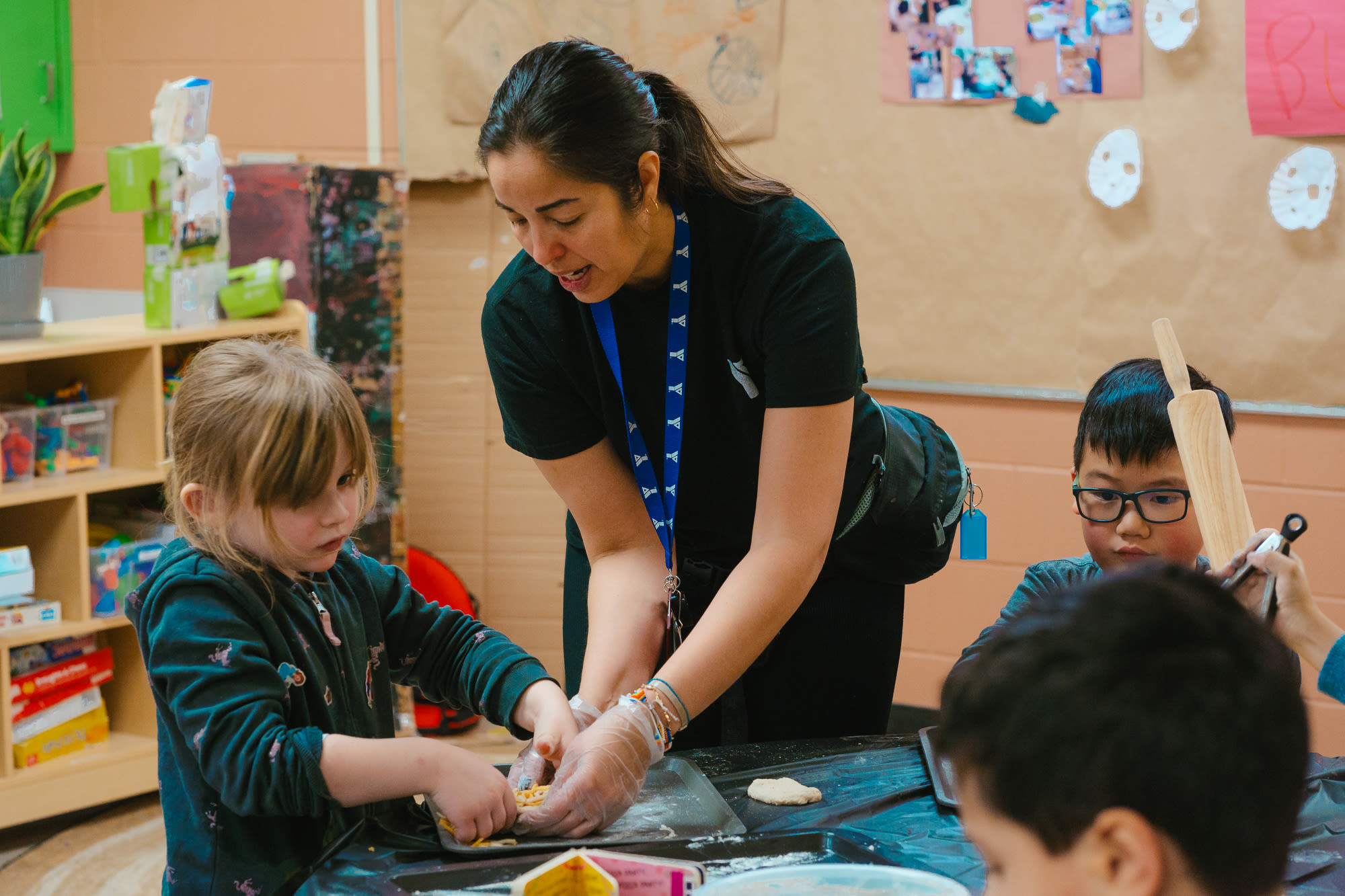 An educator in a black YMCA t-shirt assists a child with a pizza-making activity in a child care classroom.