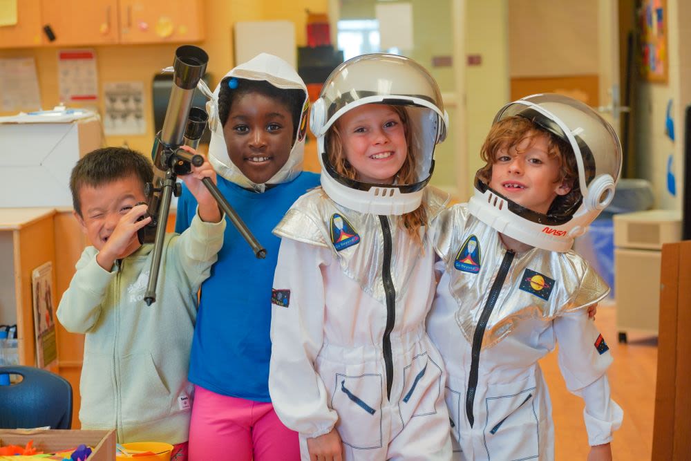 Four children wearing space costumes smile in a YMCA Before & After school classroom. Two wear spacesuits, one holds a telescope, and one wears only a space helmet.