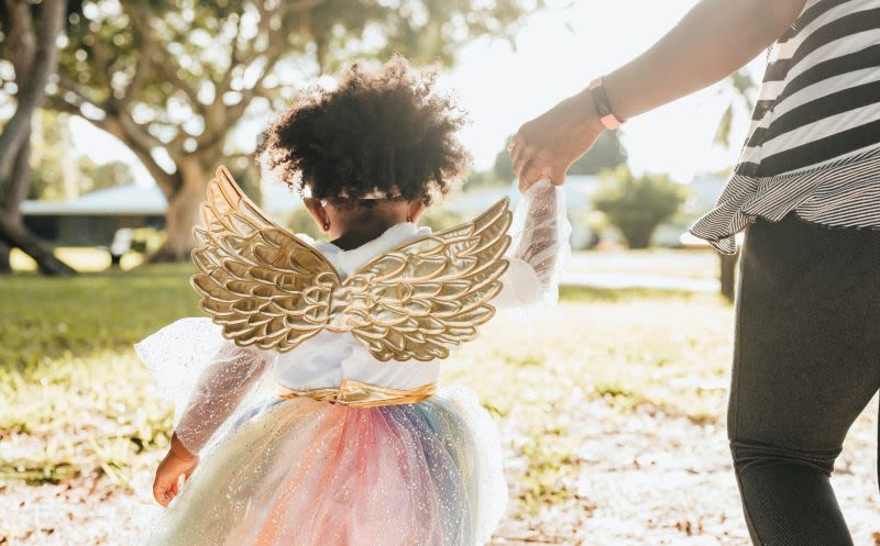 A parent holds the hand of a child in a rainbow tutu and gold wings on the sidewalk.