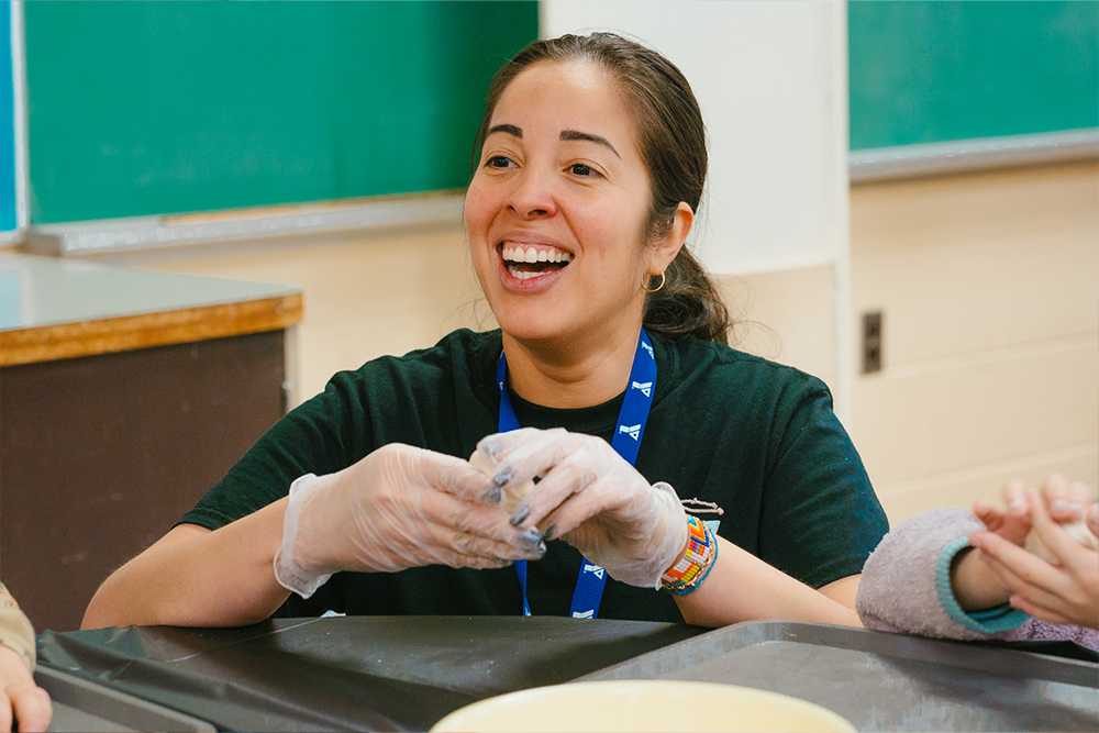 An educator in a black YMCA T-shirt smiles brightly while rolling pizza dough during a pizza-making activity with children in a YMCA child care classroom.
