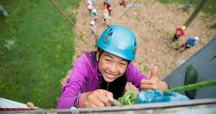  A child, clad in a purple jacket and sporting a blue helmet, ascends an outdoor rock climbing wall as part of a YMCA Summer Camp program.