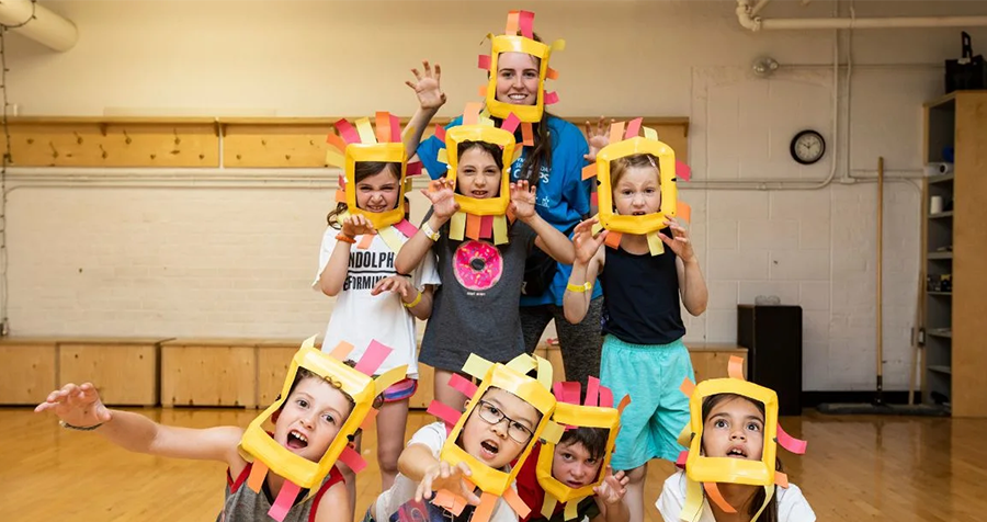  An educator and a group of children wear handmade, vibrant yellow, orange, and red lion's mane headpieces as part of an indoor activity at the YMCA Summer Camp.