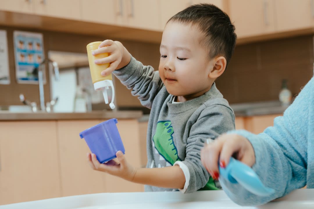 A child in a grey long-sleeve plays in a sandbox sensory box with an educator beside them.