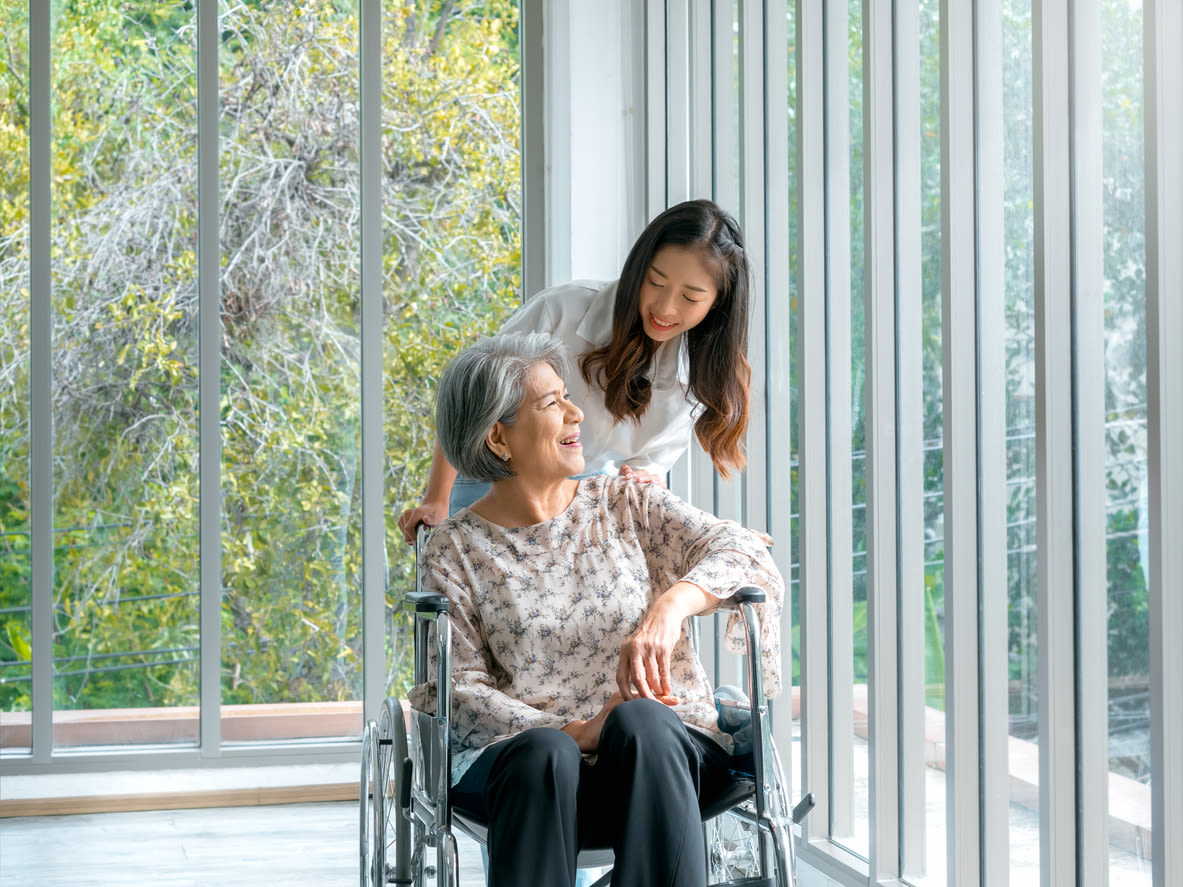 A young person with long brown hair in a white top rests a hand on an elderly person's wheelchair as they both look out at trees through large windows.