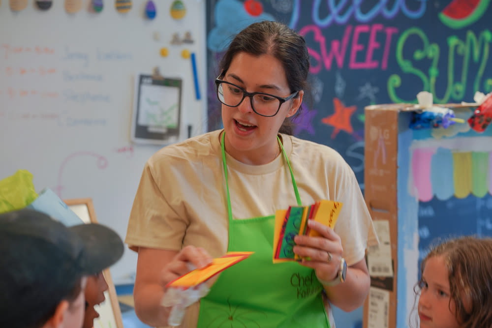 An educator in a beige top and green apron shows colourful play money to three children in a vibrant YMCA child care classroom.