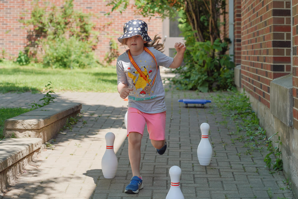 A child in a black hat with daisies, a grey graphic cartoon t-shirt, and pink shorts runs down a ramp at a Y child care centre during an obstacle course activity with bowling pins and a sitting scooter board.