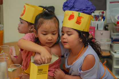 Three children dressed as chefs with yellow construction paper hats, two children select straws to place in cups for the classroom cafe.