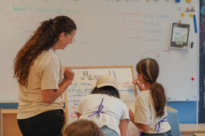 Two children write on an easel whiteboard with the classroom cafe menu notes while an educator looks over their work.