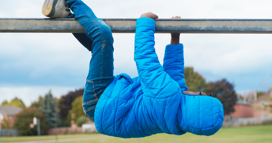 A child, dressed in a blue cold weather jacket and jeans, hangs upside down from a bar on a YMCA child care playground.
