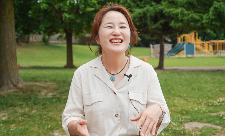 Jenny sits outdoors in front of a playground smiling brightly in a linen top.