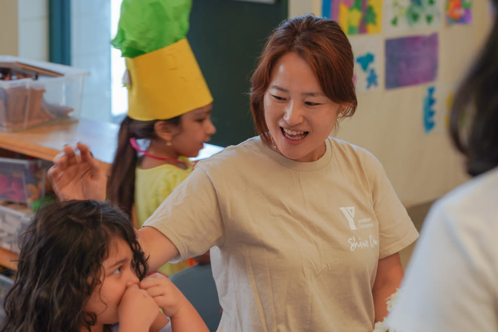 Wearing a tan YMCA Shine On logo t-shirt, Jenny sits beside a child in a licensed summer break child care classroom, smiling warmly.