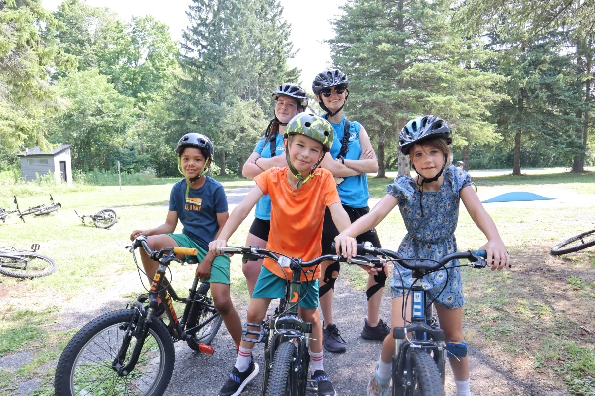 Two YMCA camp counsellors in blue t-shirts and helmets pose behind three helmeted children on bikes outdoors.