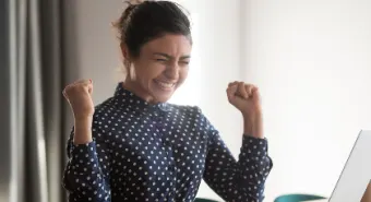 person raises arms in celebration after looking at a laptop monitor