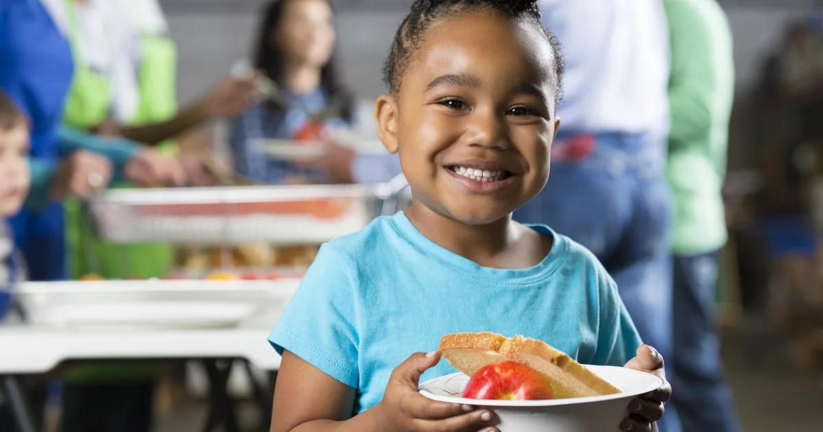 A child sadly stares out the window as she's seated in front of an empty plate on the table.