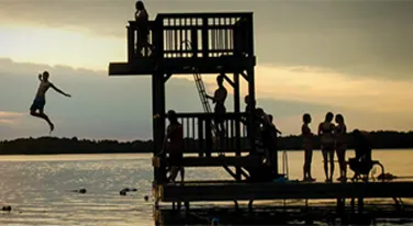 a silhouette of a group of kids jumping off a diving board in the lake