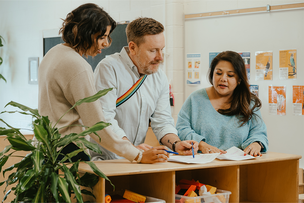 Three educators review a document while standing behind an activity cubby shelf in a YMCA child care classroom, holding pens.