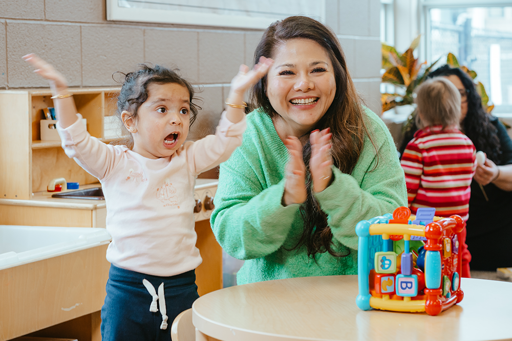 An educator in a green sweater applauds with a smile while a child next to them, wearing a pink long-sleeved shirt, excitedly waves their hands in the air while playing with a vibrant activity cube toy.