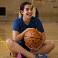 Maya sitting on the Mississauga YMCA gym floor holding a basketball and smiling