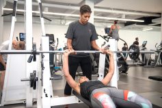 Person using a barbell to bench press and trainer coaching 