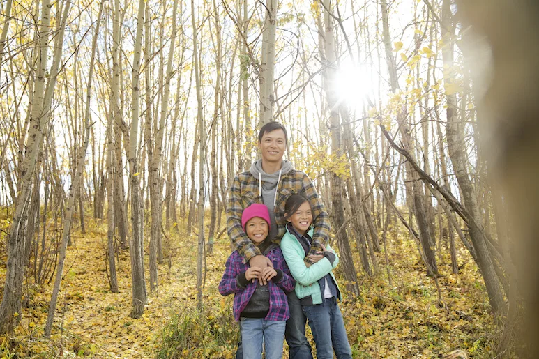 A family of two girls and one adult smiling at the camera in the woods
